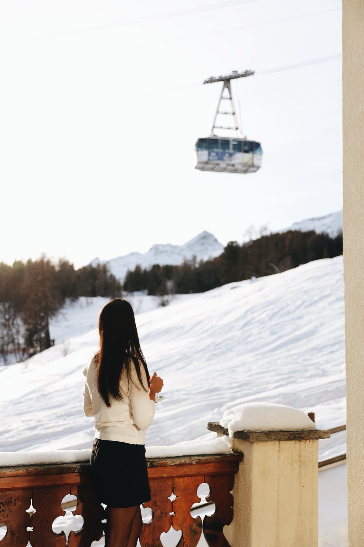 A guest on a chalet balcony above the snow, a cable car crossing beyond.